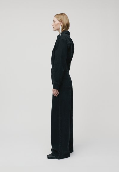 Zwarte denim jumpsuit met een aansluitend bovenstuk, wijde benen en lange mouwen. Opvallende details zijn grote oorbellen en een minimalistisch ontwerp.