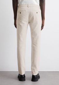 Beige linen trousers with a straight fit, two back pockets featuring gold-tone buttons, and cuffed hems. Worn with black footwear.