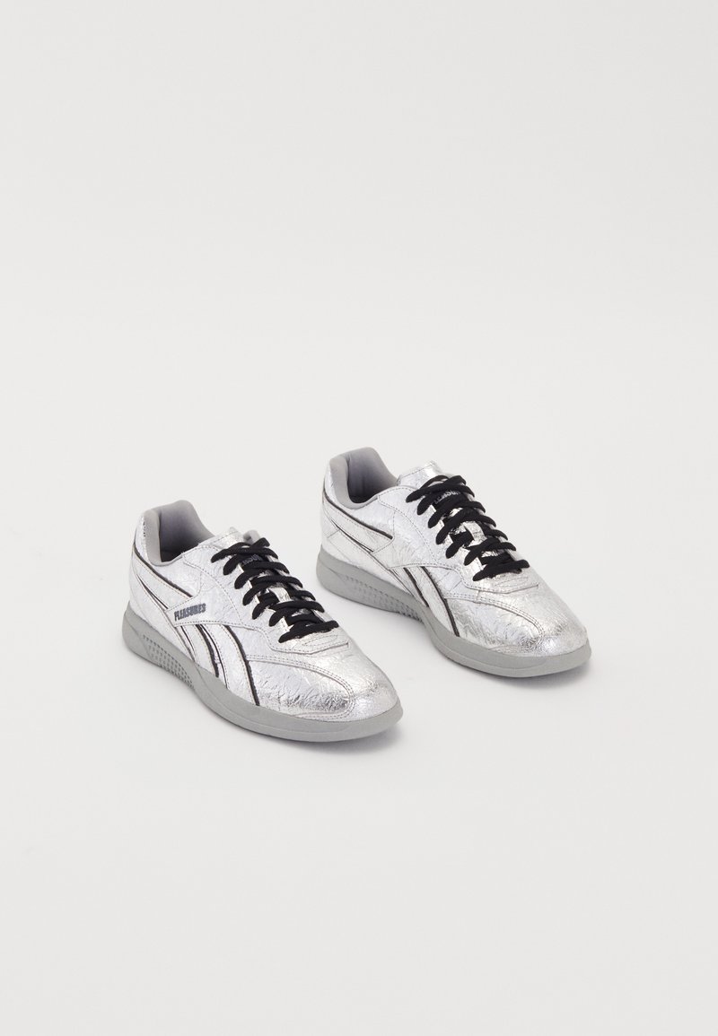 Srebrni športni copati z teksturiranim metalnim zaključkom, črnimi vezalkami, podloženim ovratnikom in sivo gumijasto podplatom. Imajo podrobnost logotipa na strani.