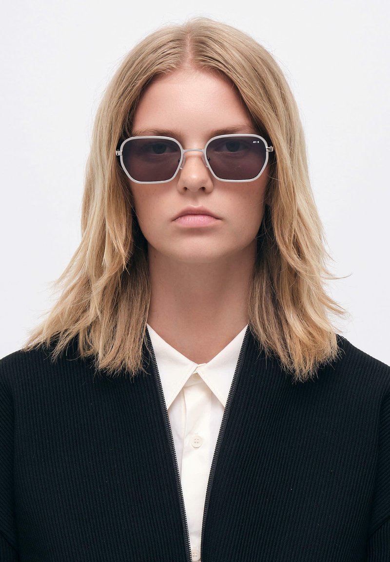 Aurinkoesterit, joissa on suorakulmainen kehys, harmaat linssit ja metalliset yksityiskohdat. Mallilla on päällään musta ribattupintainen takki valkoisen kauluspuseron päällä.