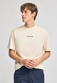 Camiseta beige clara de manga corta, con cuello redondo y texto negro "POLO CLUB" en el pecho. Material de algodón suave.
