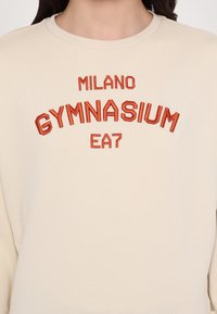 Kreminis džemperis su raudonais siuvinėtais užrašais: "MILANO GYMNASIUM EA7." Paprastas, apvaliu kaklu dizainas su lygia tekstūra.