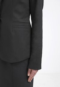 Svart skräddarsydd kavaj med en strukturerad design. Har långa ärmar, en ficka på vänster sida och en slät tygtextur.