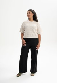 Korte mouw, beige gebreide top met horizontale strepen, gecombineerd met zwarte wijde broek en groene slidesandalen. Losse pasvorm en een gestructureerde stof.