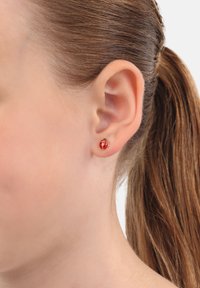 Orecchino a perno a forma di coccinella rossa con accenti dorati, caratterizzato da macchie nere e una forma arrotondata, indossato in un primo piano di un orecchio.