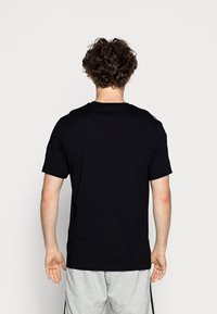 Camiseta de algodón negra con mangas cortas y cuello redondo clásico, vista desde la parte trasera. Los pantalones cortos grises son parcialmente visibles debajo.