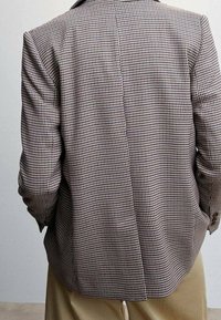 Blazer a cuadros marrón y crema con cuello con muesca, una abertura trasera y mangas con botones, confeccionado en tela texturizada.