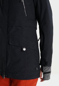 Zwarte waterdichte jas met meerdere zakken, een ritssluiting op de borst en elastische mouwen. Heeft een matte afwerking en een verstelbare zoom.