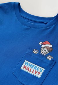 Zils T-krekls ar kabatas dizainu, kurā redzams karikatūras tēls, kas izlien no kabatas, valkājot brilles un sarkanu un baltu cepuri, ar tekstu "KUR IR VĀLIS?".