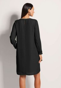 Schwarzes Kleid mit langen, gefalteten Ärmeln und rundem Ausschnitt. Schlüsselaugen-Detail auf der Rückseite und gerader Saum. Glatte Textur.