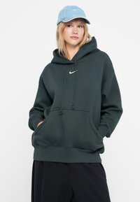 Tamsiai žalia hoodie su priekine kengūros kišene, reguliuojamu raišteliu ir baltu logotipu; derinama su šviesiai mėlynu kepurėliu. Lygus audinio paviršius.