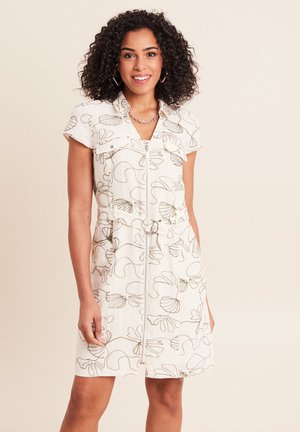 Femme aux cheveux bouclés souriante, portant une robe à manches courtes ceinturée avec une fermeture éclair et des motifs abstraits en forme de coquillage.