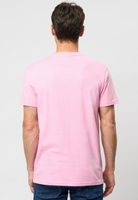 Man die van achteren is gezien, gekleed in een effen lichtroze T-shirt met korte mouwen en een blauwe spijkerbroek, tegen een neutrale achtergrond.