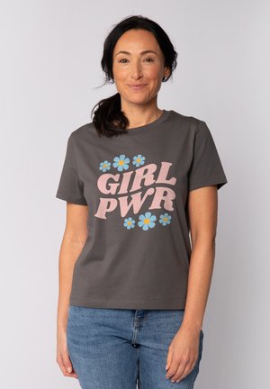 Femme aux cheveux foncés attachés en queue de cheval, portant un t-shirt gris avec le texte rose « GIRL PWR » et des fleurs bleues, associé à un jean bleu, souriant doucement.