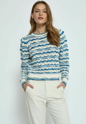 Jersey de punto con patrones en ondas horizontales azul y crema, con puños y dobladillo de canalé. Combinado con pantalones blancos.