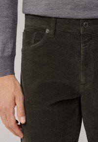 Donkergroene corduroy broek met een gestructureerd oppervlak, voorzien van zijzakken en een metalen knoop in de taille, gedragen met een grijs trui.