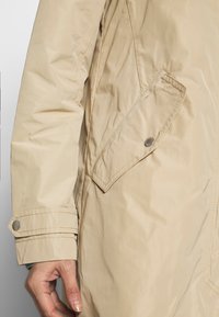 Veste imperméable beige avec une fermeture éclair et des pressions, dotée d'une poche latérale et de détails de poignets boutonnés. Texture lisse avec un léger éclat.