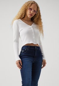 Baltas siaurintais ilgomis rankovėmis trumpas viršus su V formos iškirpte ir sagų detalėmis, derinamas su tamsiai mėlynais aukšto liemens džinsais.