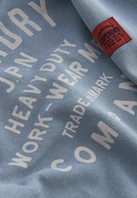 Un tessuto di colore blu chiaro mostra del testo bianco con frasi come "ABBIGLIAMENTO DA LAVORO PESANTE". È attaccato al materiale un'etichetta rossa con la scritta "SUPERDRY".