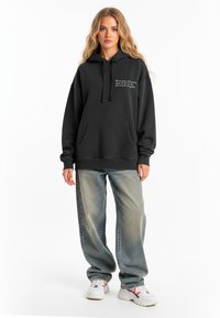 Felpa nera con tasca a marsupio, cappuccio con cordino e dettagli in bianco sul petto. Abbinata a jeans a gamba larga in lavaggio chiaro e sneakers.