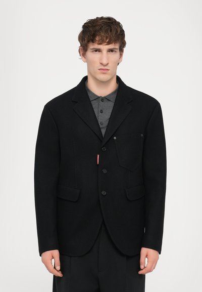 Blazer in lana nera con rever a punta, tre bottoni, due tasche frontali e una piccola etichetta con logo sul lato sinistro. Tessuto morbido.