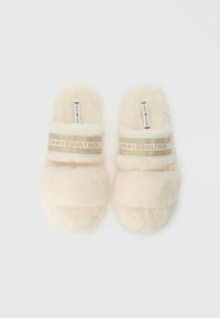 Crèmekleurige, harige slippers met brede banden en een gouden, gestructureerde band met het label "TOMMY HILFIGER." Zachte, pluche textuur overal.