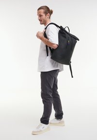 Musta käärittävä reppu vetoketjullisella etutaskulla, valmistettu sileästä materiaalista, jossa on säädettävät hihnat ja värikäs yksityiskohta vetoketjussa.