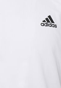 Bílý sportovní triko s texturovanou tkaninou, s černým logem Adidas a třemi černými pruhy na levém prsu.