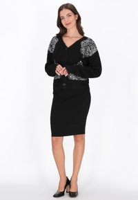 Fekete kardigán díszes fehér virágmintákkal, V nyakkivágással, hosszú ujjakkal és gombos záródással, fekete ceruzaszoknyával és magassarkú cipővel párosítva.