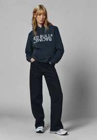 Sudadera negra con el texto en blanco "RED BULL RACING", combinada con pantalones negros holgados y zapatillas blancas. Los materiales parecen suaves y casuales.