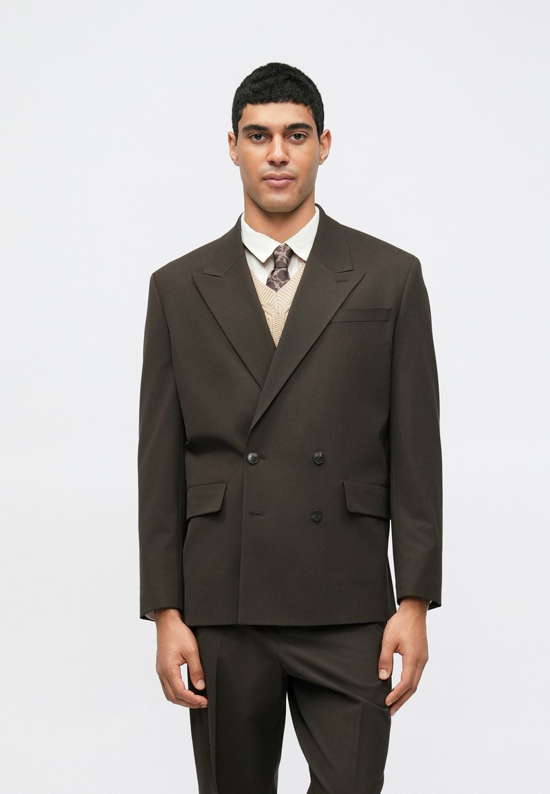 Σκούρο καφέ σακάκι κομμάτι με δύο κουμπιά, με δύο μπροστινές τσέπες, φορεμένο πάνω από ανοιχτόχρωμο γιλέκο και γραβάτα. Λείο ύφασμα με δομημένα ώμους.