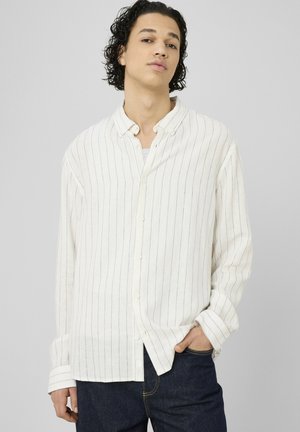 Jeune homme aux cheveux bouclés portant une chemise blanche à manches longues avec de fines rayures verticales et un jean bleu foncé, main dans la poche.