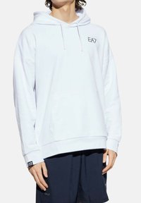 Personne portant un sweat à capuche blanc EA7 avec poche avant et shorts bleu marine, se tenant devant un fond clair et uni.