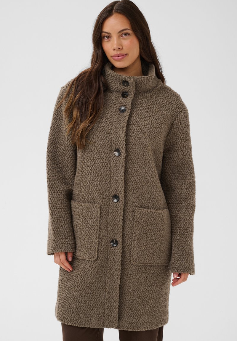 Femme portant un manteau marron texturé mi-longueur avec de grandes poches avant et des boutons noirs, debout devant un fond clair uni.