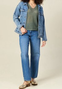 Veste en denim de couleur bleu clair avec des poches à rabat, associée à un haut en maille vert en V, un jean droit bleu et des chaussures métalliques.