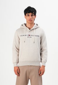 Man in een beige Tommy Hilfiger hoodie met kangoeroezak en beige geribbelde broek, staand tegen een witte achtergrond.
