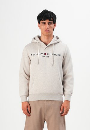 Uomo che indossa una felpa con cappuccio beige di Tommy Hilfiger con tasca a marsupio e pantaloni a coste beige, in piedi davanti a uno sfondo bianco.