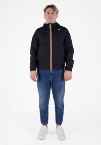 Giacca nera con zip e polsini elastici, caratterizzata da un logo bianco e una striscia gialla lungo la zip. Indossata sopra jeans di denim blu e sneakers bianche.