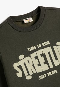 Ciemnozielony bawełniany sweatshirt z okrągłym dekoltem, z białym napisem "CZAS NA JAZDĘ STREETLIFE PO PROSTU JEŹDŹ." Miękka tekstura.