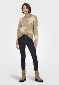 Beige trui met een roomkleurig sterpatroon, ronde halslijn en ribbels aan de mouwen. Gecombineerd met donkerblauwe skinny jeans en tan enkellaarsjes met ritsen.