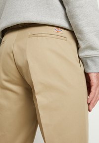 Pantalon de travail beige avec poche passepoilée arrière et écusson au logo Dickies porté avec un sweat-shirt gris clair, montrant le bas du torse et la main.