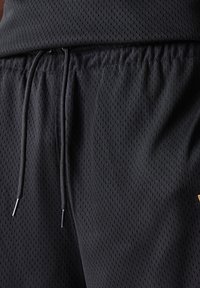 Svart treningsshorts laget av et strukturert og pustende mesh-stoff, med elastisk midjebånd og snøring, som har metallisk spisser.