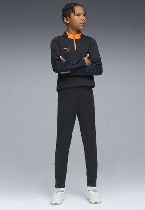 Conjunto deportivo negro con diseño de media cremallera, con acentos naranjas y mangas texturizadas, combinado con zapatillas deportivas blancas.
