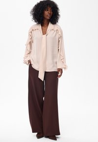 Jasnoróżowa bluzka z falbanami i wiązanym kołnierzykiem, zestawiona z luźnymi brązowymi spodniami. Gładka tekstura materiału, szerokie rękawy i luźny krój.