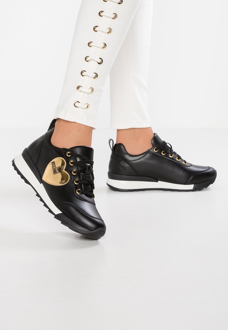 Black leather sneakers featuring gold heart detail, gold eyelets, black laces, and a white rubber sole with a textured grip.
