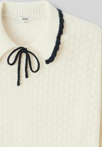 Pull en tricot blanc avec un motif texturé, col en côtes et bordure festonnée bleu marine. Présente un nœud noué au cou. Étiquette de marque : BZB.
