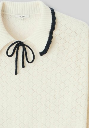 Pull en tricot blanc avec un motif texturé, col en côtes et bordure festonnée bleu marine. Présente un nœud noué au cou. Étiquette de marque : BZB.