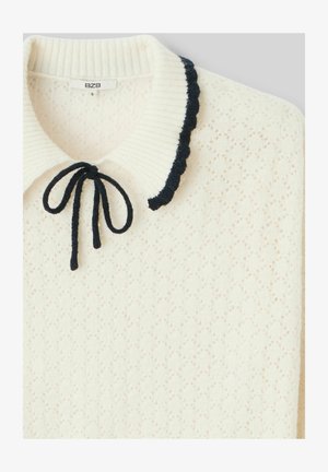 Pull en tricot blanc avec un motif texturé, col en côtes et bordure festonnée bleu marine. Présente un nœud noué au cou. Étiquette de marque : BZB.