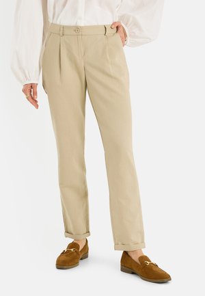 Pantalones beige a medida con dobladillos puño, combinados con mocasines de ante marrones y una blusa blanca con mangas abullonadas.