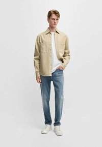 Beige lange mouwen shirt over een witte t-shirt, gecombineerd met lichtblauwe jeans en witte sneakers, met een relaxte pasvorm en twee borstzakken.